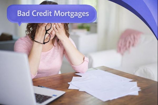 A stressed woman sits at a wooden table with paperwork spread out beside a laptop, covering her face with her hands while holding a pair of glasses. A blue speech bubble in Open Sans reads “Bad Credit Mortgages,” with curved blue Connect Mortgages brand accents in the top-right corner.