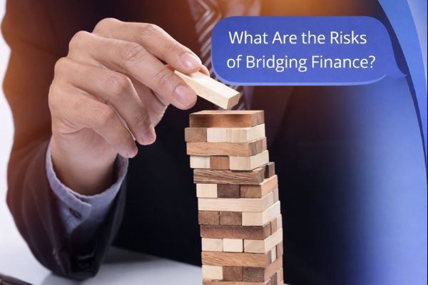 A 600×300 promotional graphic shows a close-up of a man in a suit carefully placing a wooden block on top of a Jenga-style tower. On the right, Connect Mortgages’ curved blue brand accents frame a large blue speech bubble containing the title, “What Are the Risks of Bridging Finance?”.