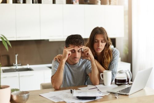 Adverse Credit Mortgage: Stressed couple at a kitchen table reviewing bills and paperwork, with a laptop and coffee on the table as the woman comforts the man holding his head. T