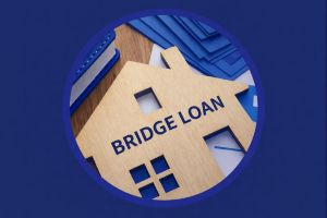 Circular badge showing a wooden house cutout with the words “BRIDGE LOAN” in white Open Sans on a dark blue strip. A dark blue notebook and light blue papers sit in the background, with a thick dark blue circular border around the image.