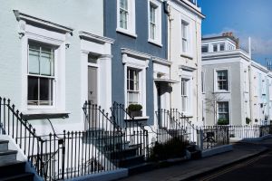 Row of pastel-coloured Victorian terraced houses with white-framed windows, black iron railings, and front steps along a quiet residential street on a sunny day.