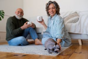 Smiling older couple relaxing at home with mugs of tea or coffee, sitting on a rug beside the bed while the woman pets a hairless cat in the foreground thinking about equity release mortgages