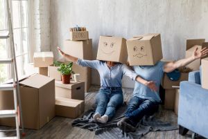 Two people sitting on the floor in a new living room with cardboard boxes around them, wearing boxes on their heads and celebrating after sorting paperwork for moving home mortgages.