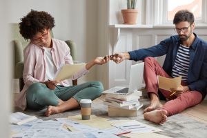 Couple sitting on the floor at home reviewing paperwork and documents with a laptop and coffee, discussing finances and exploring a Second Charge Mortgage as an option to raise additional funds.