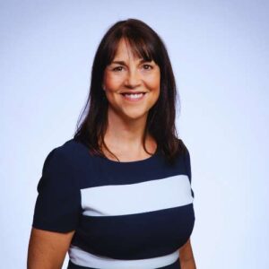 Meet Liz Syms: A smiling woman with dark shoulder-length hair and bangs stands against a light background, wearing a navy dress with a wide white horizontal stripe across the chest.